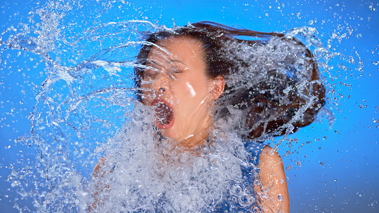 Surprise young woman being splashed with water against blue background.