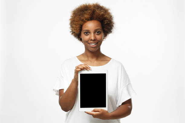 mujer afroamericana joven aislada sobre fondo gris que presenta tablet en blanco vertical de la pantalla a los espectadores con sonrisa, copyspace para publicidad de productos o aplicaciones - black sign holding vertical fotografías e imágenes de stock