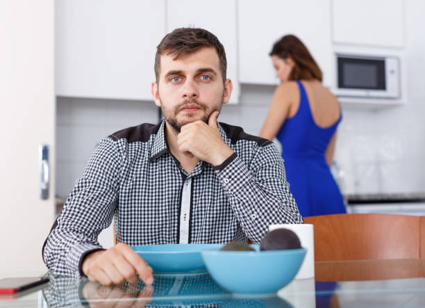 istock Young male sitting at kitchen table while woman preparing food 1026693424