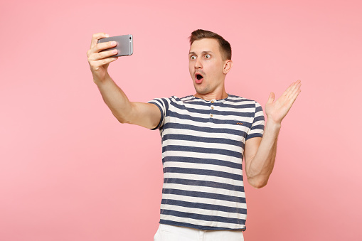 Portrait of smiling young man wearing striped t-shirt doing selfie shot on modern mobile phone isolated on trending pastel pink background. People sincere emotions lifestyle concept. Advertising area