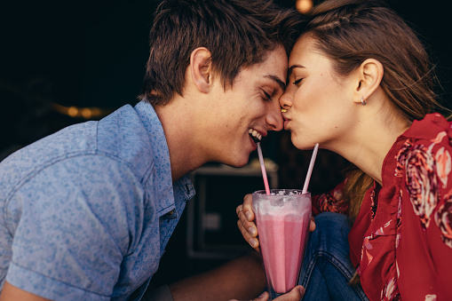 Woman kissing her boyfriend on his nose while on date at a restaurant. Couple at a restaurant in romantic mood sharing a milkshake.