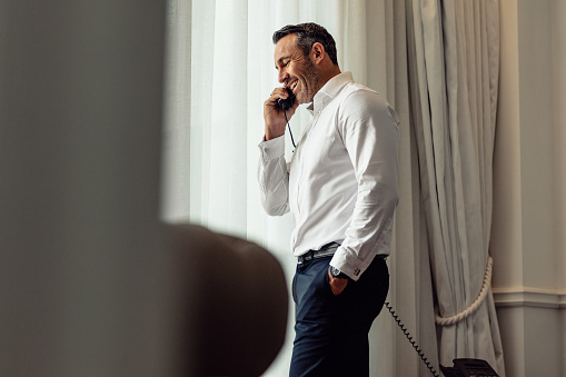 Businessman talking on phone while standing in a hotel room. CEO on business trip staying on a hotel and making phone call from landline phone.