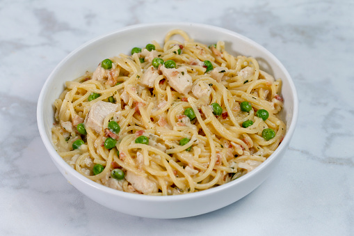 A bowl of pasta with grilled chicken, bacon bits and peas in a creamy carbonara sauce on a marble counter.