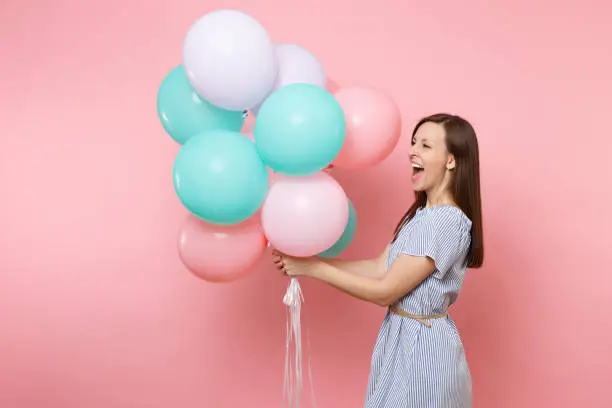 Portrait of joyful laughing young happy woman wearing blue dress holding colorful air balloons looking aside isolated on bright pink background. Birthday holiday party, people sincere emotion concept