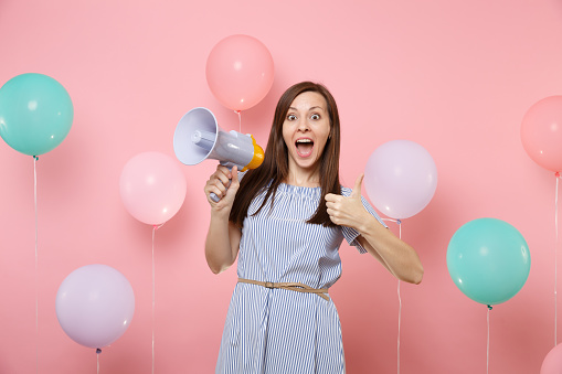 Portrait of excited young attaractive woman wearing blue dress holding megaphone showing thumb up on pink background with colorful air baloons. Birthday holiday party, people sincere emotions concept