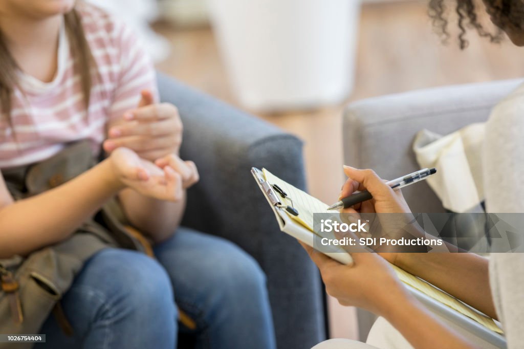 Mental health professional takes notes during session Unrecognizable teenage girl shares her troubles with a female mental health professional. The mental health professional is taking notes. Mental Health Professional Stock Photo