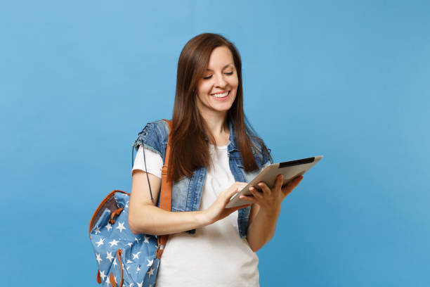 istock Young pleasant beautiful woman student in denim clothes with backpack holding and using working on tablet pc computer isolated on blue background. Education in college. Copy space for advertisement. 1026760362