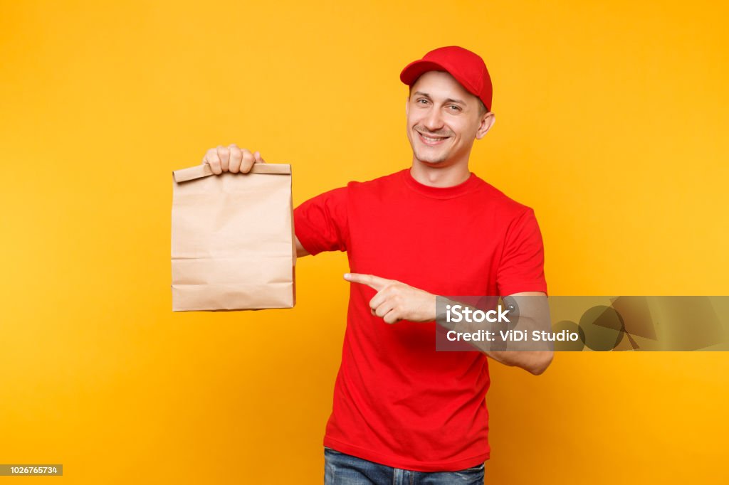 Man in red cap, t-shirt giving fast food order isolated on yellow background. Male employee courier hold empty paper packet with food. Products delivery from shop or restaurant to home. Copy space. Man in red cap, t-shirt giving fast food order isolated on yellow background. Male employee courier hold empty paper packet with food. Products delivery from shop or restaurant to home. Copy space Delivering Stock Photo