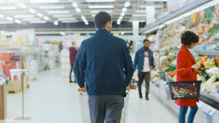 At the Supermarket: Man Pushing Shopping Cart Through Fresh Produce Section of the Store. Store with Many Customers Shopping. Following Back View Shot.