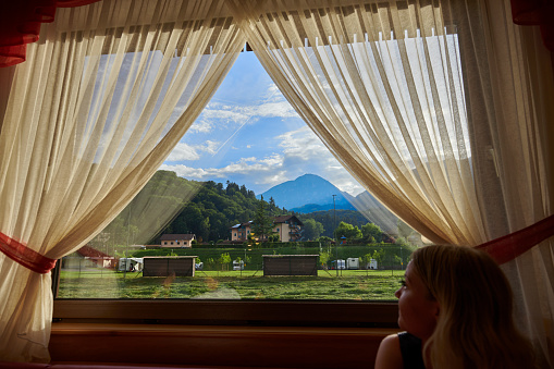Woman looking at Italian landscape through window with curtain.