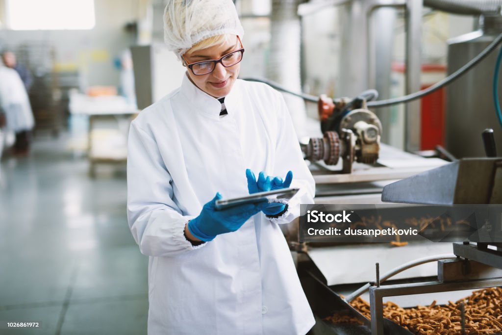 Joven trabajadora con ropa estéril es comprobar la calidad de productos en fábrica de alimentos. - Foto de stock de Alimento libre de derechos