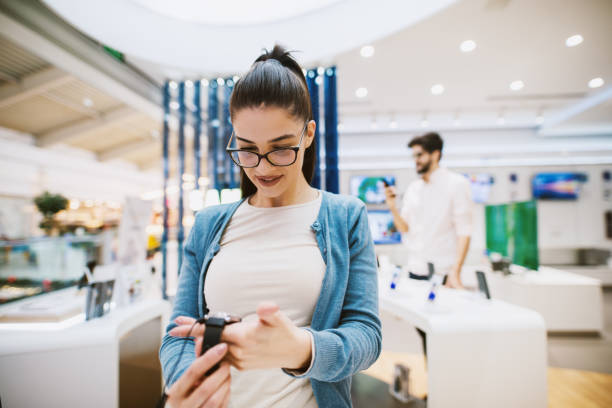 istock Beautiful modern girl is testing new telephone in tech store while her boyfriend is looking around in a store. 1026871282