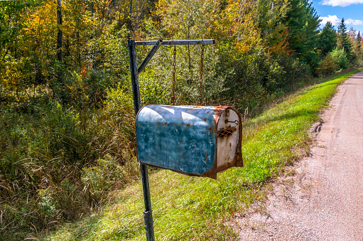 Weathered rural mailbox hanging from chains & metal post at roadside on an bright sunny autumn day. 3/4 view at eye level