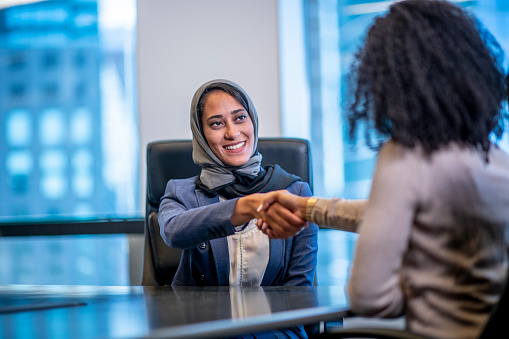 A woman wearing a head scarf is shaking hands with another woman after making a deal. They are indoors in an office building.
