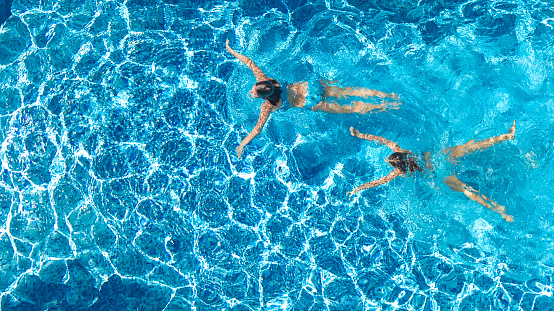 Aerial top view of girls in swimming pool water from above, active children swim, kids have fun on tropical family vacation, holiday resort concept