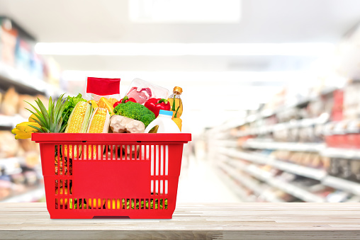 Food and groceries in red shopping basket on wood table with blurred suppermarket aisle in background