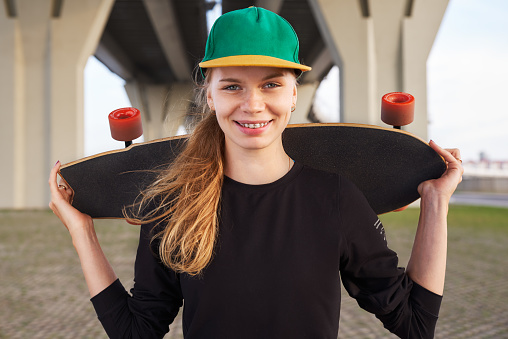 The girl looks at the camera. The skateboarder keeps a longboard on his shoulders and wearing a fashionable cap.