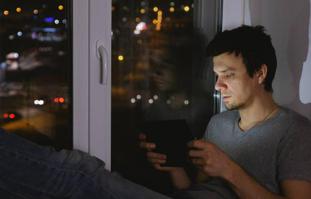 istock Man brunette playing game on tablet computer. Sitting on the windowsill in the dark. 1027059498