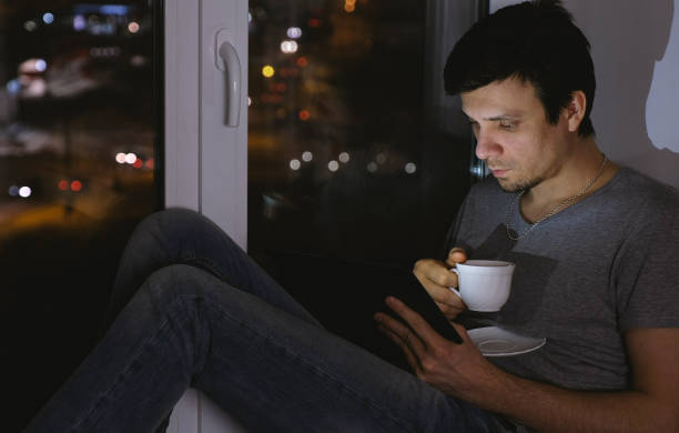 istock Man reading something on tablet and drinking tea. Sit on the windowsill in the dark at night. 1027059600