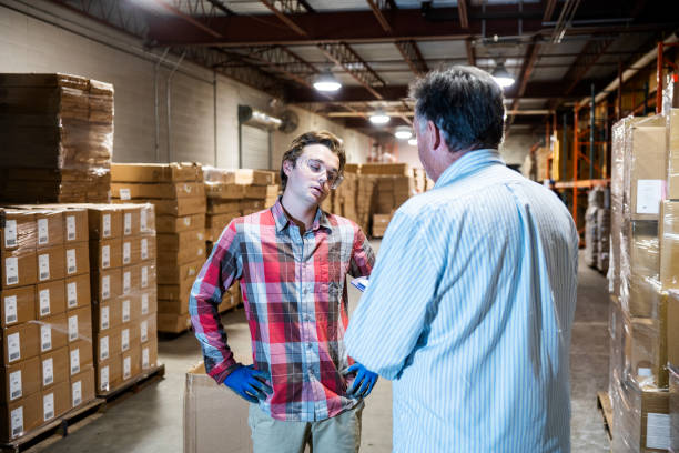 A warehouse manager talks with a younger warehouse worker. A mature warehouse manager discusses issues with a younger male warehouse worker. The young worker has a look of disagreement and annoyance. The manager is raising job performance or safety issues with the young man. fired employee stock pictures, royalty-free photos & images