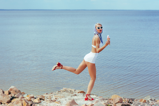 young woman in retro swimsuit holding ice cream and posing on rocky beach