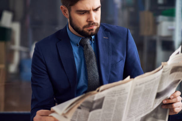 Displeased frowning male entrepreneur with beard concentrated on article reading newspaper in airport Displeased entrepreneur reading newspaper newspaper airport reading business person stock pictures, royalty-free photos & images