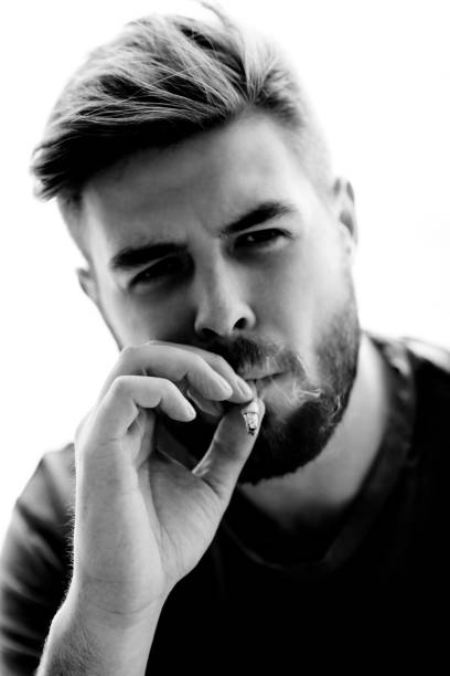 istock Serious brutal handsome young man with beard taking drag on cigarette while smoking, black and white portrait 1027122210