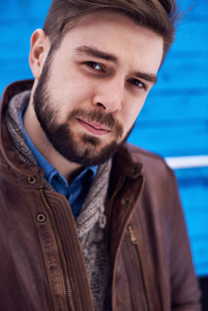 istock Content handsome young man in brown leather jacket and sweater looking at camera and standing outdoors 1027124858