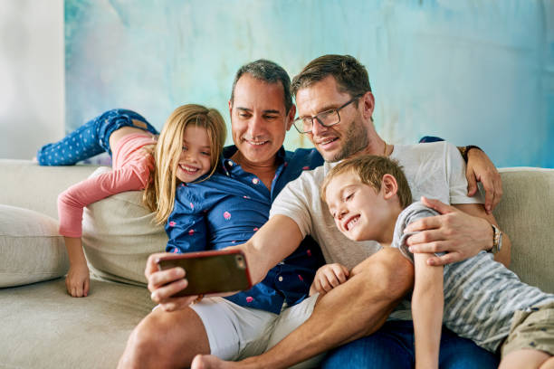Get in here, we're taking a selfie Cropped shot of an affectionate family of four taking selfies on the sofa at home gay daddy stock pictures, royalty-free photos & images