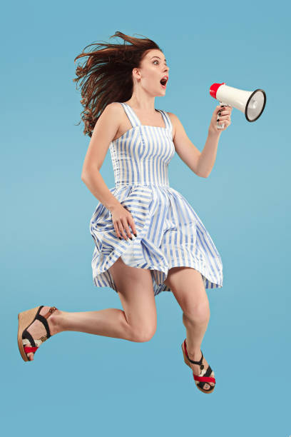 istock Beautiful young woman jumping with megaphone isolated over blue background 1027169070
