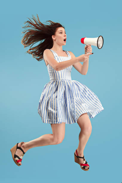 istock Beautiful young woman jumping with megaphone isolated over blue background 1027169248