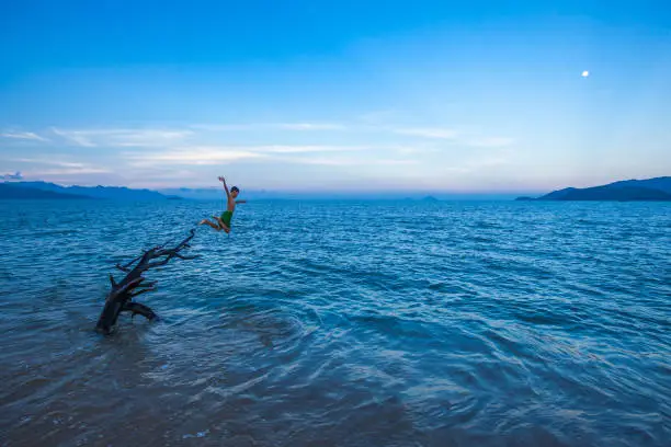 Child jumps off driftwood into blue sea at dusk, Nha Trang, Vietnam