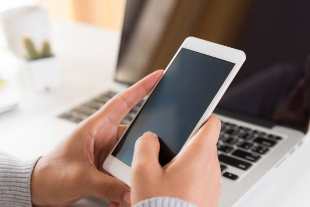istock Woman hand holding white mobile phone on a table with a laptop in office. 1027194934
