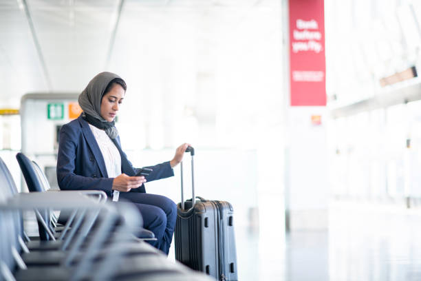 Ready To Travel A Muslim woman is sitting in an airport. She is holding her suitcase and checking her smartphone. airports canada stock pictures, royalty-free photos & images