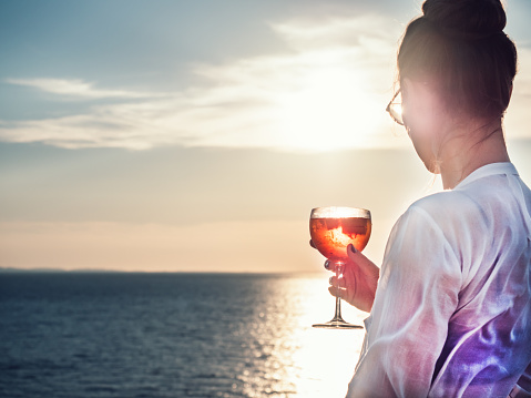 Young woman in glasses with a glass of beautiful, pink cocktail in hand on an open deck of a cruise ship on a background of sunset and sea waves