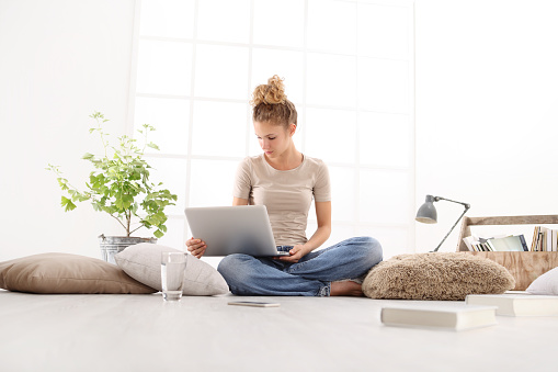 young woman with computer, smartphone and books, sitting on the floor in living room on white wide window in the background