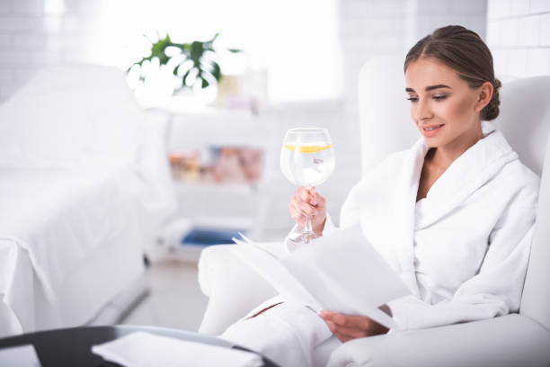 istock Smiling young lady holding glass of lemon water while reading booklet 1027355552