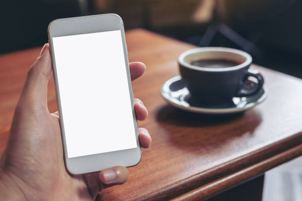 istock Mockup image of hand holding white mobile phone with blank desktop screen with coffee cup on wooden table in cafe 1027450452