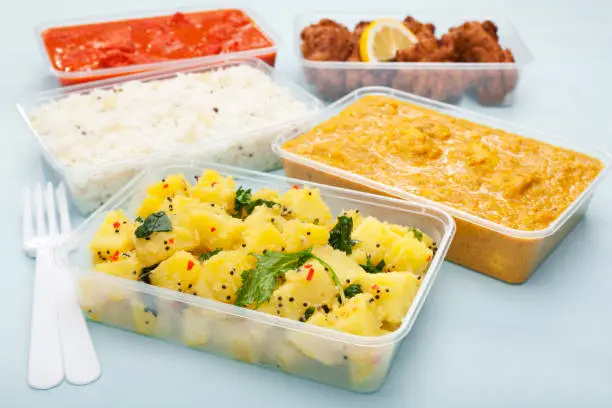 A selection of Indian takeaway food in plastic containers on a blue background. Aloo saag (potato spinach curry), chicken tikka masala, chicken bhoon or  bhuna, basmati rice and onion bhaji or pakora.