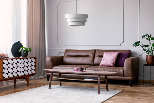 istock Real photo of light grey sitting room interior with window with drapes, leather sofa with pillows, coffee table on carpet and cupboard with decor 1027582636