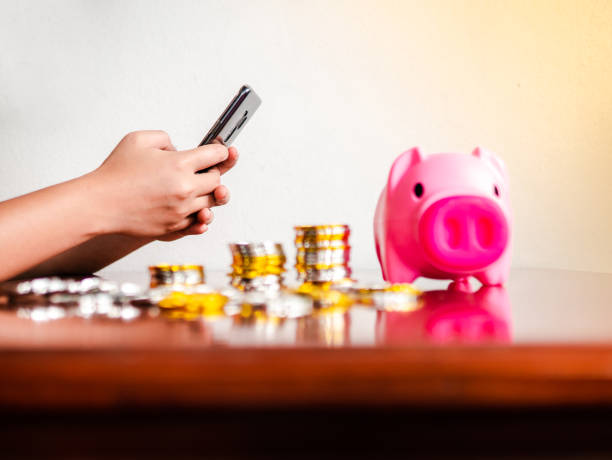 main tenant et utilisant l’app téléphone intelligent avec rouleaux de nombreuses pièces de monnaie, piggy boîte sur table en bois. représenter à économiser de l’argent, plan d’investissement, gagner sur internet, paiement ou faire du shopping  - piggy bank currency savings finance photos et images de collection