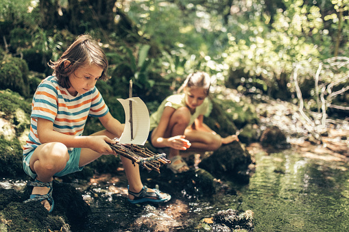 Children putting handmade raft sailboat made of wood in the river. Beautiful nature reflecting on water surface