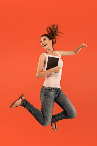 istock Image of young woman over blue background using laptop computer or tablet gadget while jumping 1027666680