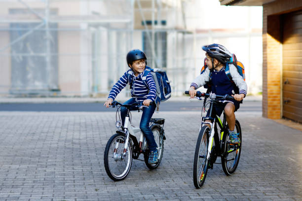 zwei school kid jungen in schutzhelm fahren mit fahrrad in der stadt mit rucksäcken. glückliche kinder in bunten kleidern, radfahren auf dem fahrrad unterwegs zur schule. sicherer schulweg für kinder im freien - people child twin smiling stock-fotos und bilder