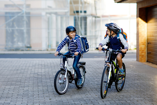 Two school kid boys in safety helmet riding with bike in the city with backpacks. Happy children in colorful clothes biking on bicycles on way to school. Safe way for kids outdoors to school.