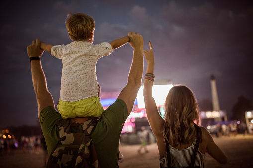 Young family with son festival or concert. Father carrying child on shoulders. Dusk time.