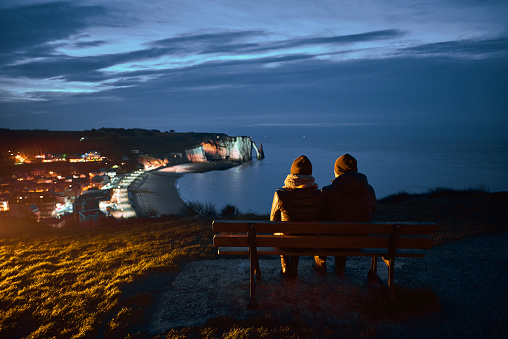 rear view of couple sitting on bench in Etretat region in night time, admiring the city.