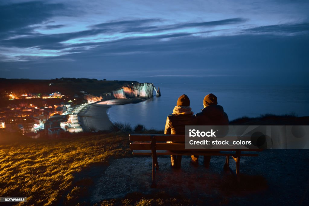 admiring amazing view rear view of couple sitting on bench in Etretat region in night time, admiring the city. Night Stock Photo