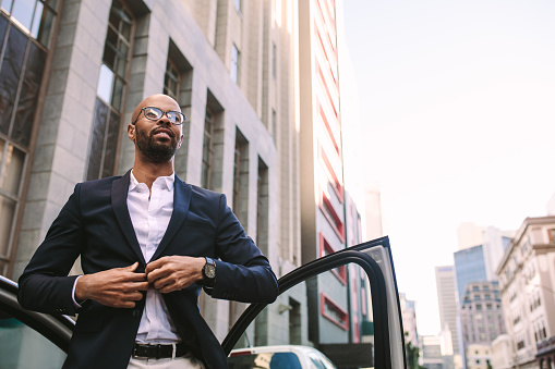 Portrait of a handsome young businessman getting down from car on road and buttoning his jacket. African man standing by his car in city.