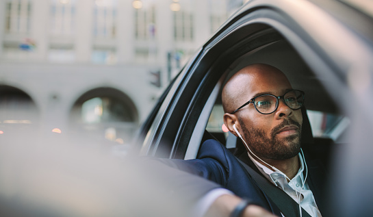 Relaxed businessman wearing earphones and listening musing while driving a car. African businessman traveling in a car through city street.